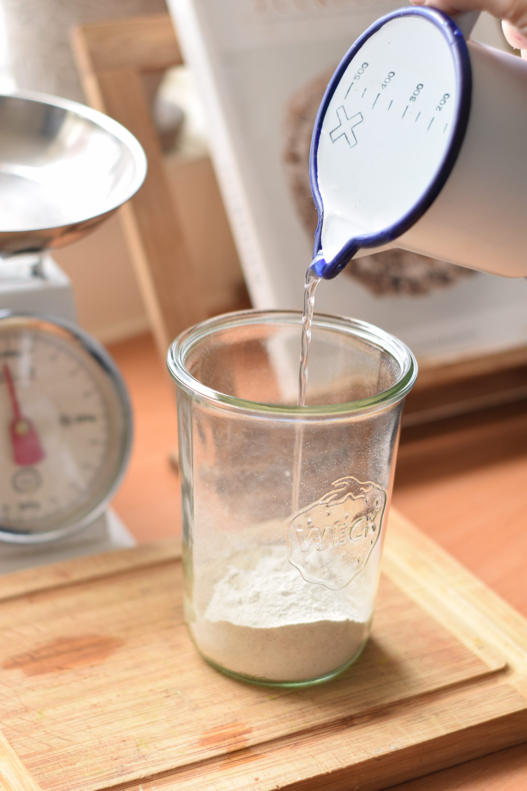 Pouring of water from an enamel measuring cup into sourdough starter in a glass jar