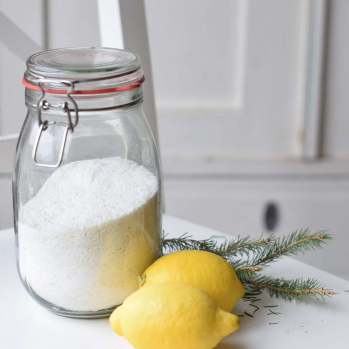 A glass jar of laundry soap with two lemons in front. It's standing on a white chair before an antique, white cabinet.