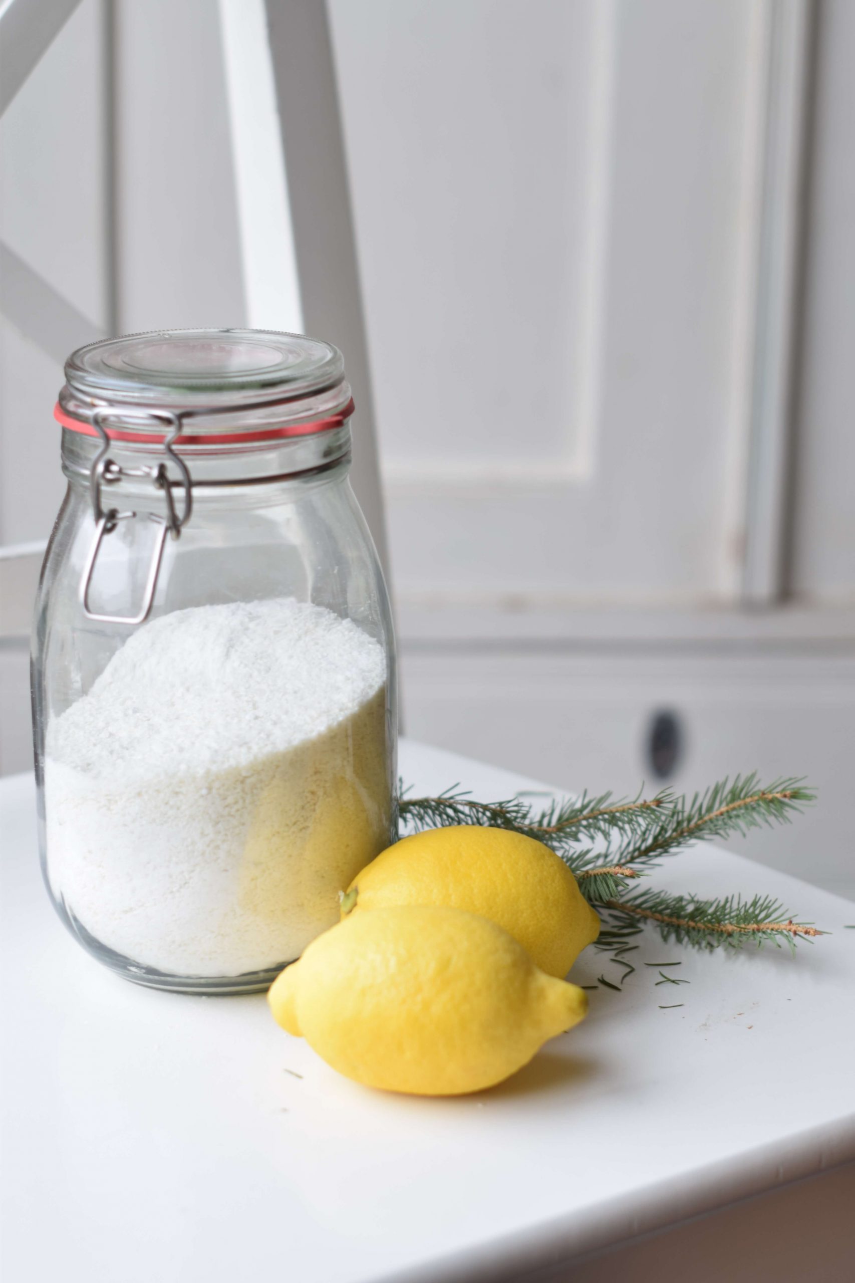 A glass jar of laundry soap with two lemons in front. It's standing on a white chair before an antique, white cabinet.