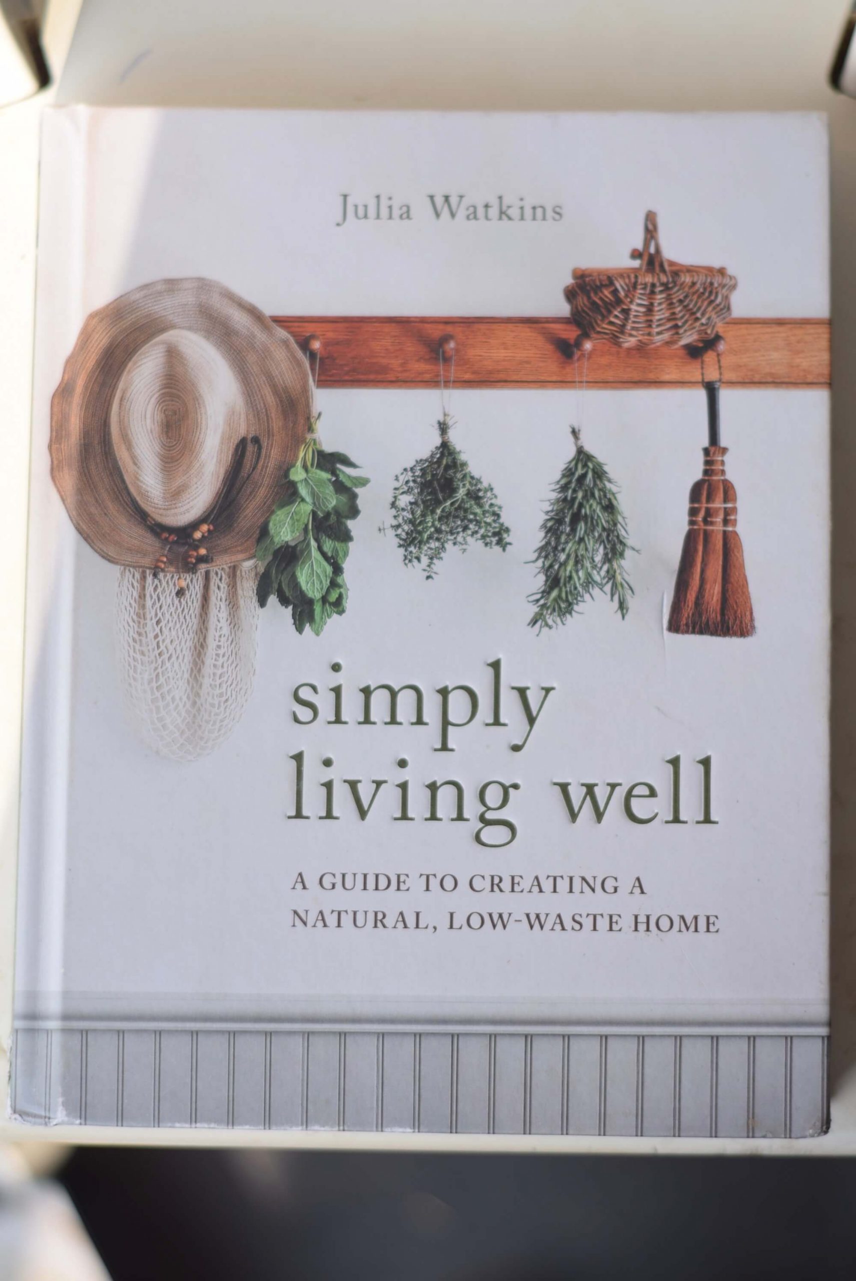 Cover of the book Simply living well by Julia Watkins. A hat, fresh herbs and a small broom hang from a coat rack with a basket on top