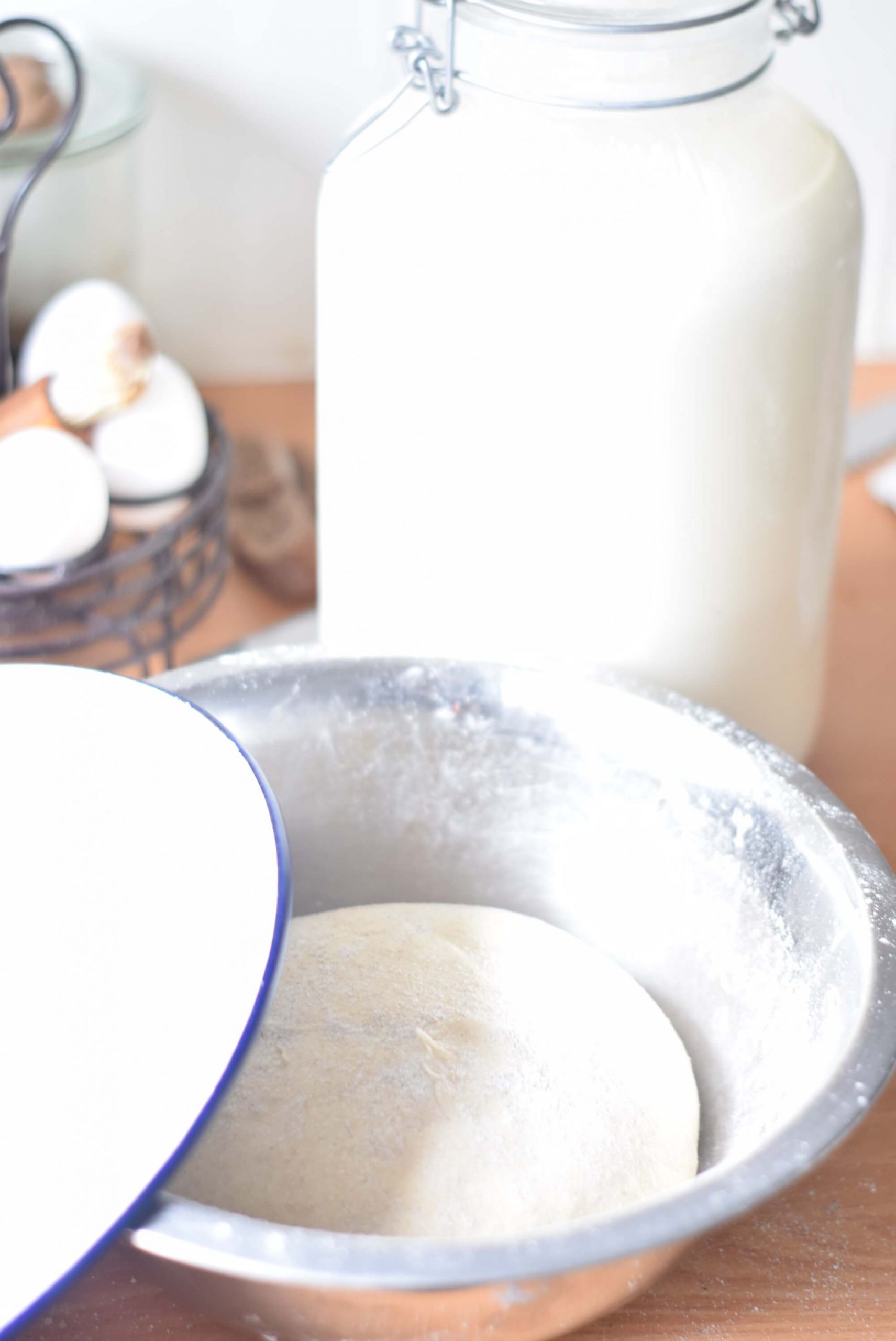 A bowl of dough for sourdough doughnuts with a large glass jar full of fresh, raw milk. An enamel plate with a blue rim covers the bowl of dough about 1/3.