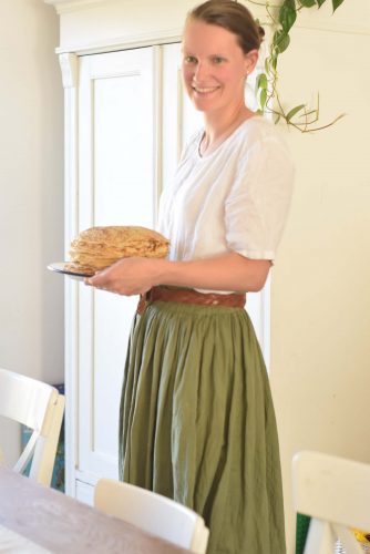 Carying a stack of einkorn spelt pancakes to the table. Wearing a long green skirt and a white linnen top with a brown leather belt.