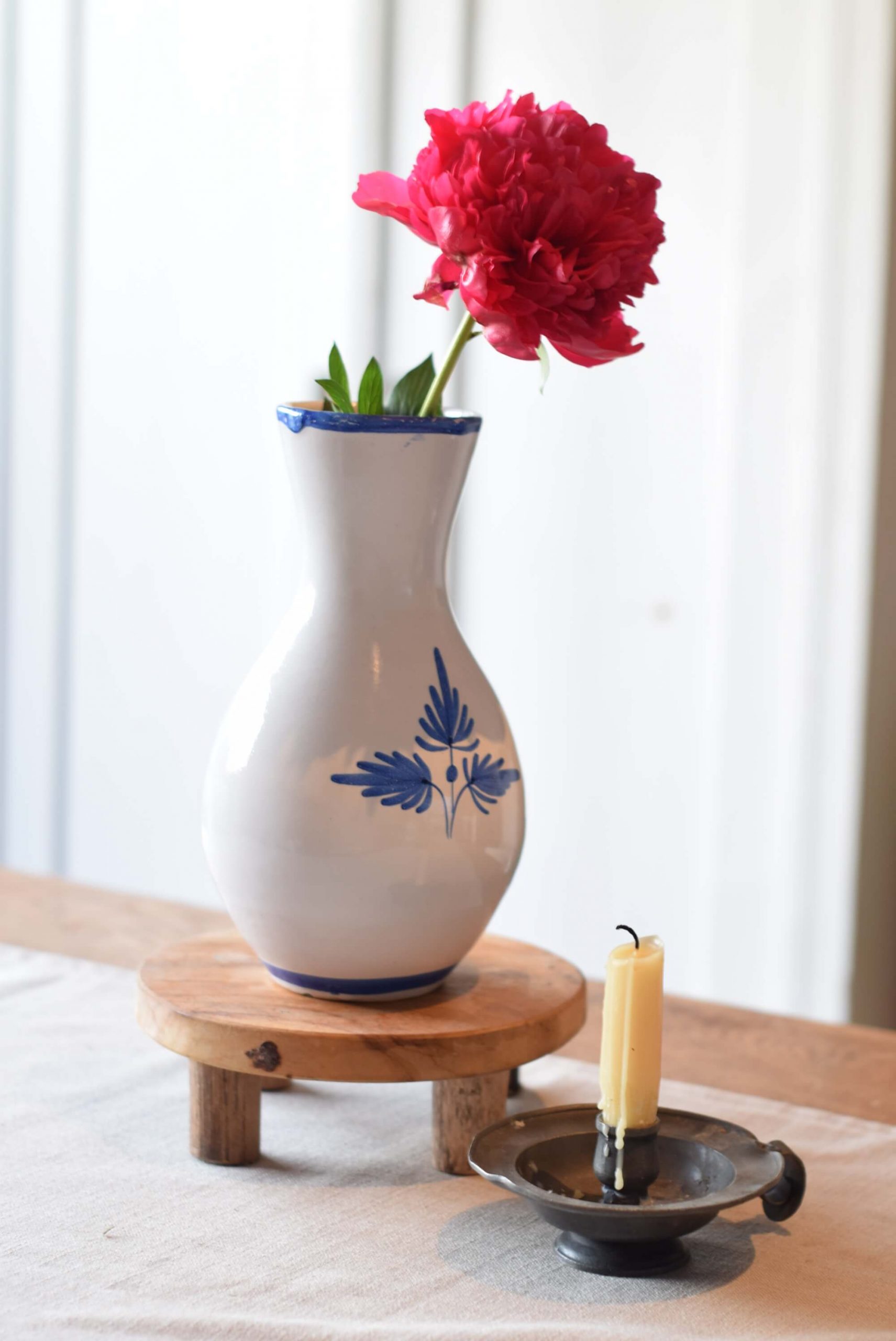 A gifted vase from the thriftstore in white with blue flowers painted on it and with a blue rim. It stands on a wooden plank and has a pink peony. Next to it stands a beeswax candle.