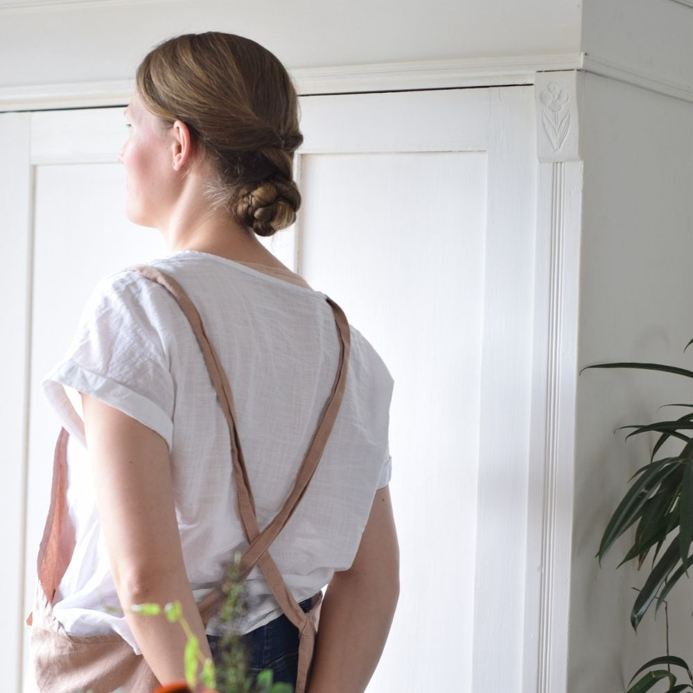 A woman wearing a pink linen apron, standing in front of a white antique cabinet, showcasing vintage homemaking skills and a simple, old-fashioned lifestyle.