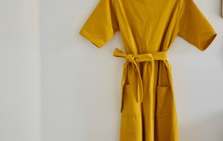 Yellow linen look dress hanging on antique hook with white wall and open door in background, next to a baby crib with a cozy blanket.