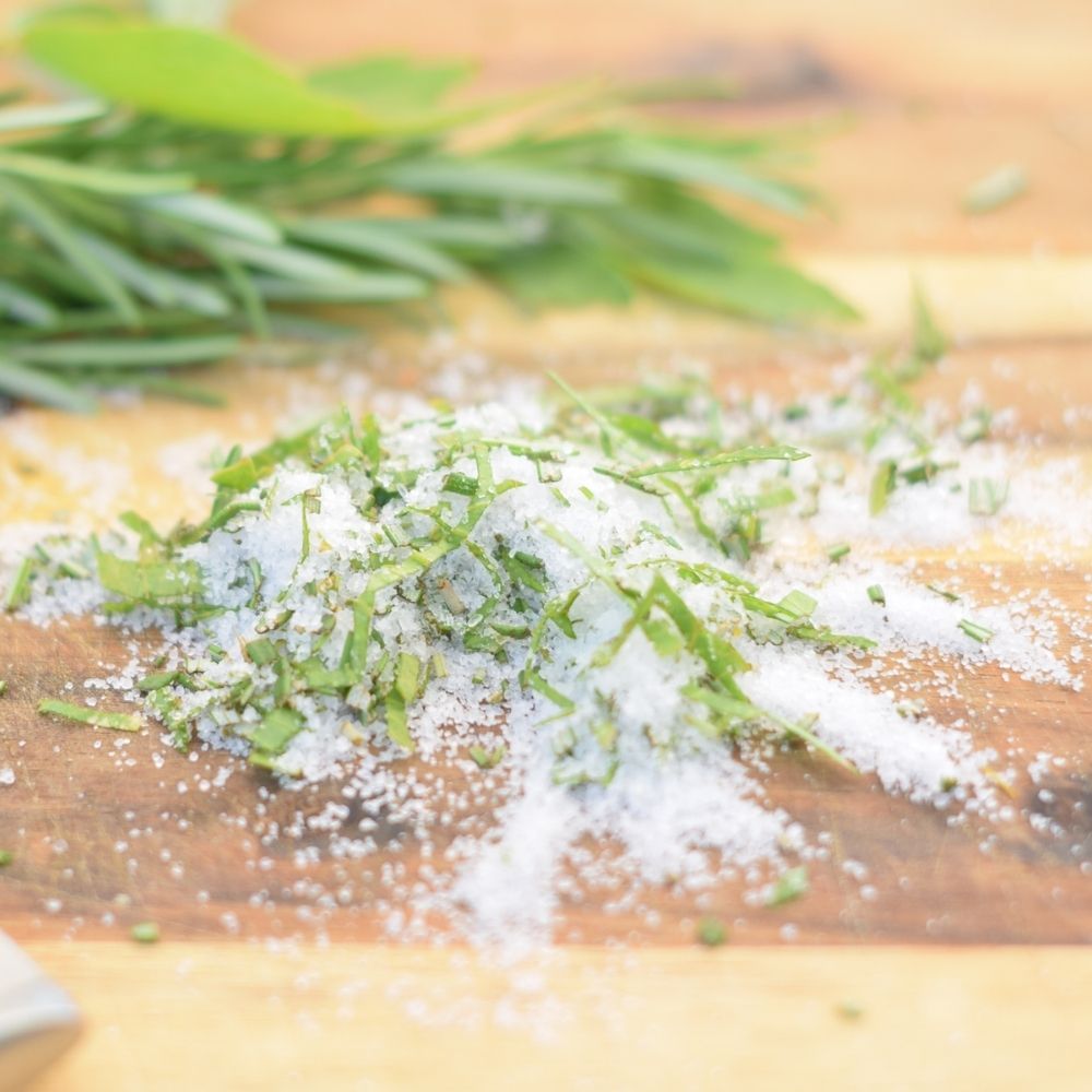 A wooden cutting board displays a blend of salt and vibrant herbs, showcasing homemade culinary herb salt ready for use.