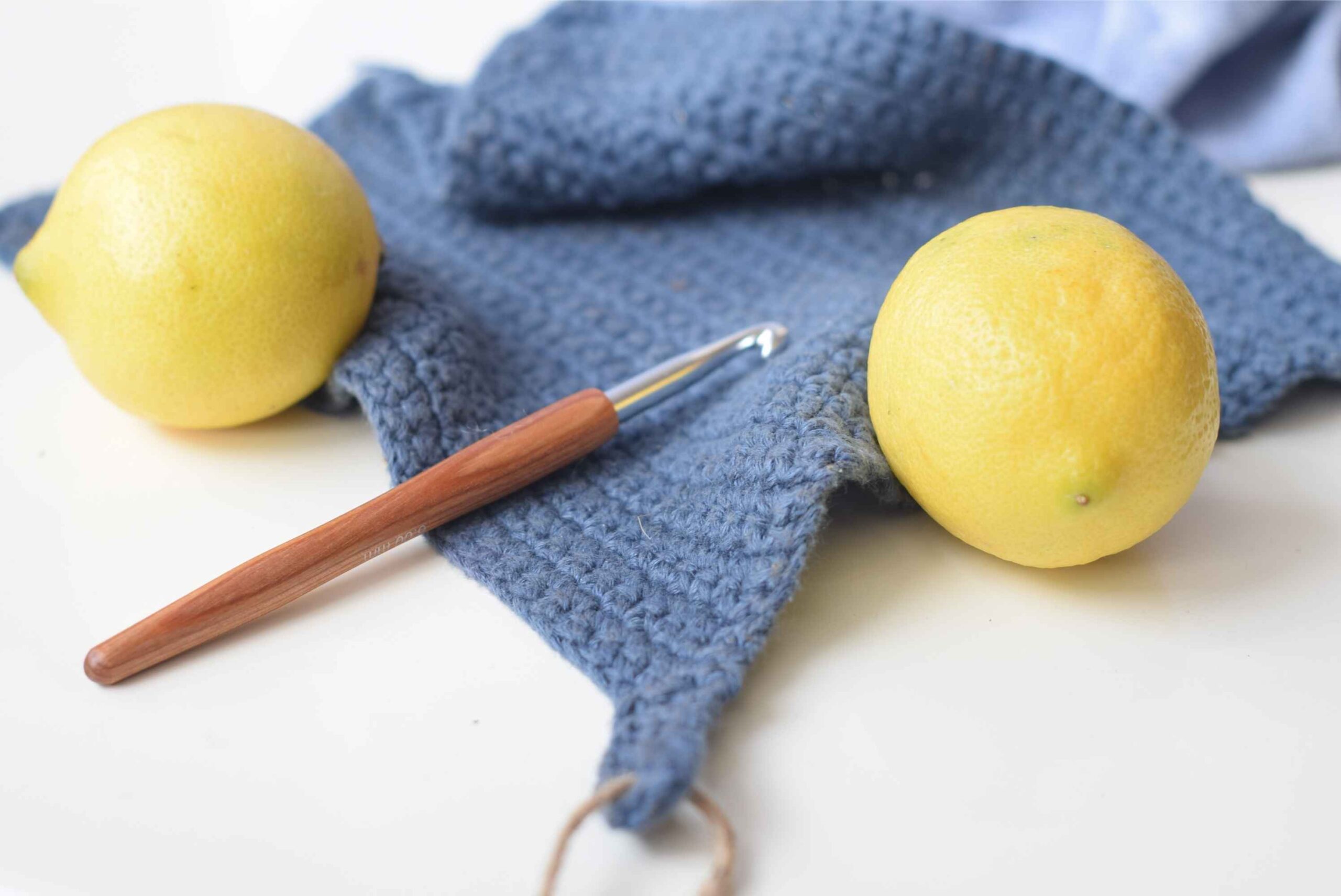 Blue crocheted dishcloth paired with bright yellow lemons and a crochet hook, displayed on a clean white surface.