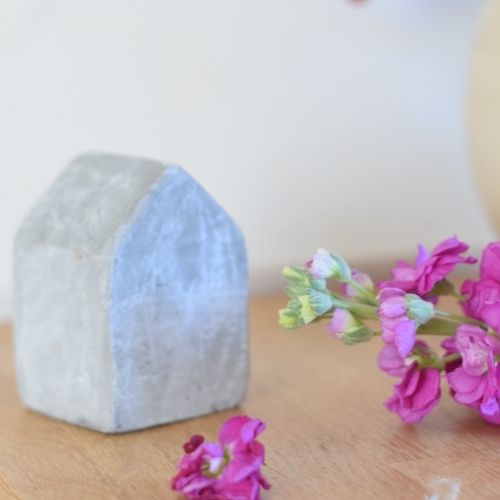 A small concrete house figurine placed on a wooden kitchen counter with a delicate purple flower beside it, representing home, comfort, and resilience.